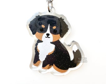 May include: A black, brown, and white Bernese Mountain Dog keychain with glitter accents. The keychain is made of clear acrylic and features a cartoon-style illustration of a dog sitting. The keychain is attached to a silver metal key ring.