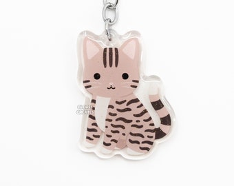May include: A silver key chain with a clear acrylic charm featuring a cartoon tabby cat with brown stripes. The cat is sitting with its tail curled around its legs. The charm has the text "GLORIE CREATES" printed on it.