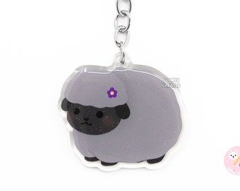 May include: A gray sheep keychain with a purple flower on its head. The sheep is made of clear acrylic with glitter embedded in the material. The keychain is attached to a silver metal ring.