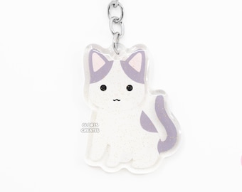Pode incluir: Um chaveiro de gato branco e roxo com glitter e um anel de chave prateado. O gato está sentado com o rabo enrolado em torno de suas pernas. O chaveiro tem o texto "CLORIS CREATES".