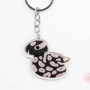 May include: A black and white snake keyring with glitter accents. The snake is curled up in a relaxed pose. The keyring is attached to a silver metal ring.