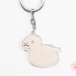 May include: A white snake with a blue eye and a pink tongue is depicted on a clear acrylic keyring. The keyring is attached to a silver metal ring and chain.