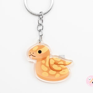 May include: A yellow and orange cartoon snake keyring with a silver chain and ring. The snake is curled up in a circle with its head sticking out. The keyring is made of clear acrylic.
