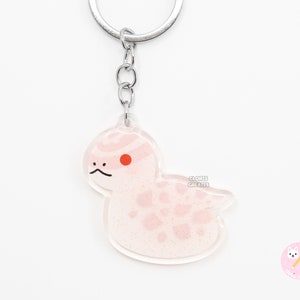 May include: A pink and white snake keychain with a red eye. The keychain is made of clear acrylic and has a silver chain.