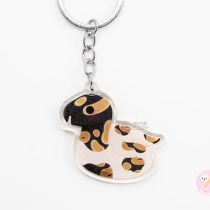 Puede incluir: Un llavero de acrílico transparente con una serpiente blanca y negra con marcas naranjas. El llavero tiene un anillo y una cadena de metal plateado. El texto "GLORIS CREATES" está impreso en el llavero.