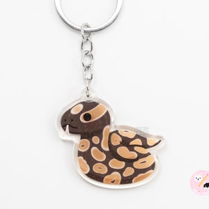 May include: A brown and tan snake with a glittery texture, curled up in a circle, is printed on a clear acrylic keyring. The keyring has a silver metal ring and chain.