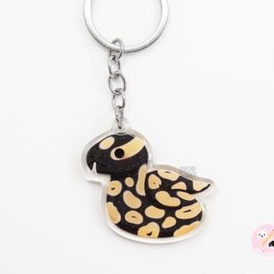 May include: A black and yellow snake keyring with a silver key ring. The snake is cartoonish and has a cute expression. The keyring is made of clear acrylic.