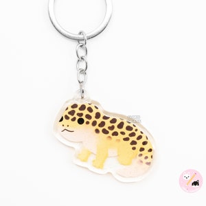 Könnte beinhalten: Ein durchsichtiger Acryl-Schlüsselanhänger mit einer Cartoon-Illustration eines Leopardgeckos. Der Gecko ist gelb mit braunen Flecken und hat einen weißen Bauch. Der Schlüsselanhänger ist an einem silbernen Metallring und einer Kette befestigt.