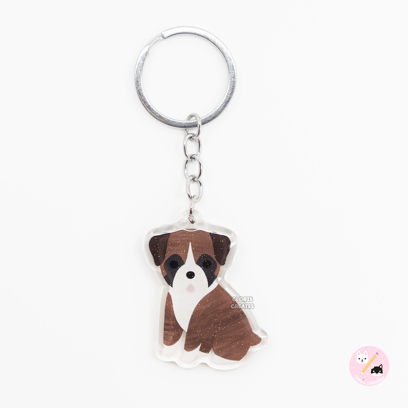 May include: A brown and white boxer dog keychain with glitter accents. The keychain is made of clear acrylic and has a silver metal ring.