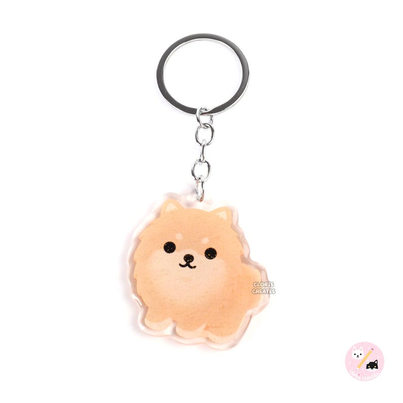 May include: A clear acrylic keychain featuring a cartoon illustration of a fluffy orange Pomeranian dog. The keychain is attached to a silver metal ring and chain.