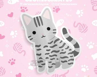 May include: A gray and white striped cat sticker with a cute cartoon face. The sticker is on a pink background with white paw prints and hearts.