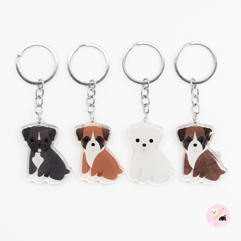 May include: Four keychains featuring different dog breeds: a black and white boxer, a brown and white boxer, a white maltese, and a brown and white boxer. Each keychain has a silver key ring.