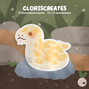 May include: A yellow and white cartoon snake with a pink tongue curled up on a grey rock. The snake is on a brown background with green leaves.