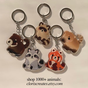 May include: Five different animal keychains with silver key rings. The keychains are made of clear acrylic and feature a raccoon, a red panda, a bear, a dog, and another raccoon. Each keychain has the text "CLORIS CREATES" printed on it.