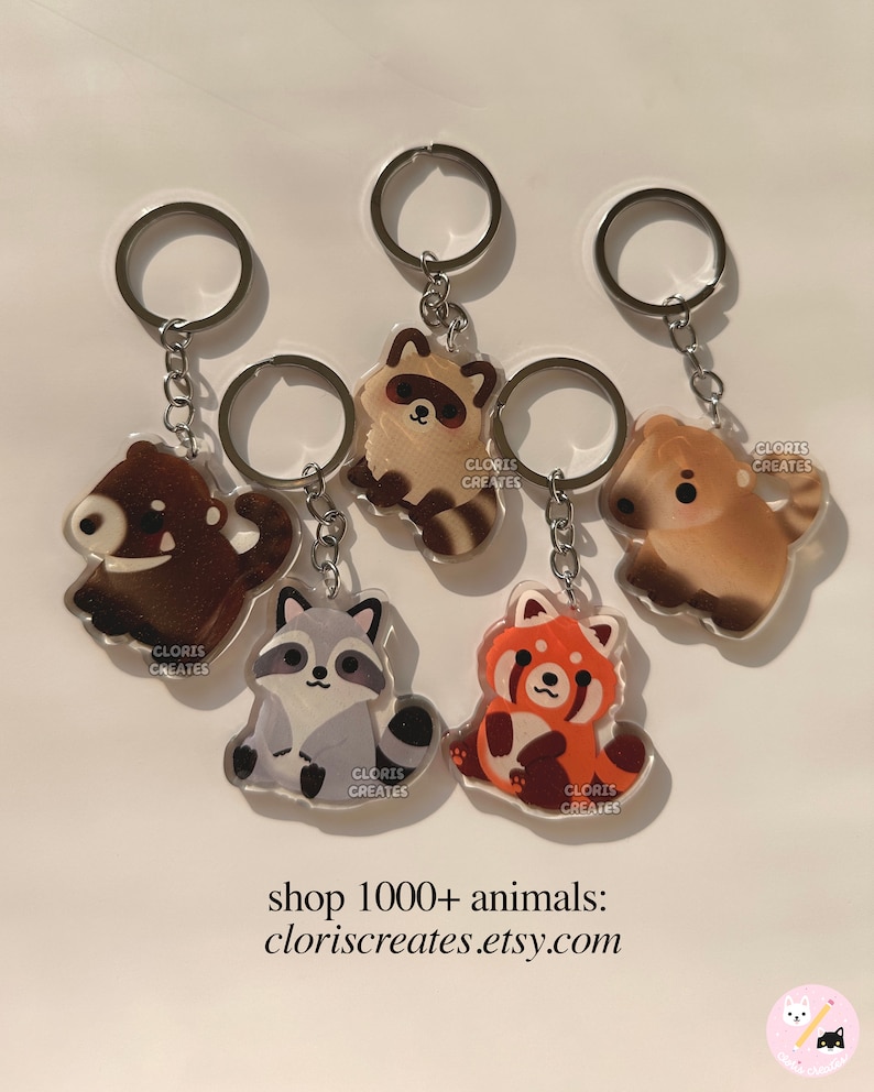 May include: Five different animal keychains with silver key rings. The keychains are made of clear acrylic and feature a raccoon, a red panda, a bear, a dog, and another raccoon. Each keychain has the text "CLORIS CREATES" printed on it.