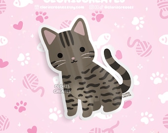 May include: A sticker featuring a cartoon tabby cat with brown and black stripes. The cat is sitting with its tail curled. The background is pink with paw prints, hearts, fish, and yarn ball illustrations. The sticker has the text "CLORISCREATES" and a website address.