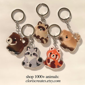 May include: Five different animal keychains with silver key rings. The keychains are made of clear acrylic and feature a raccoon, a red panda, a bear, and two different dogs. Each keychain has the text "CLORIS CREATES" printed on it.