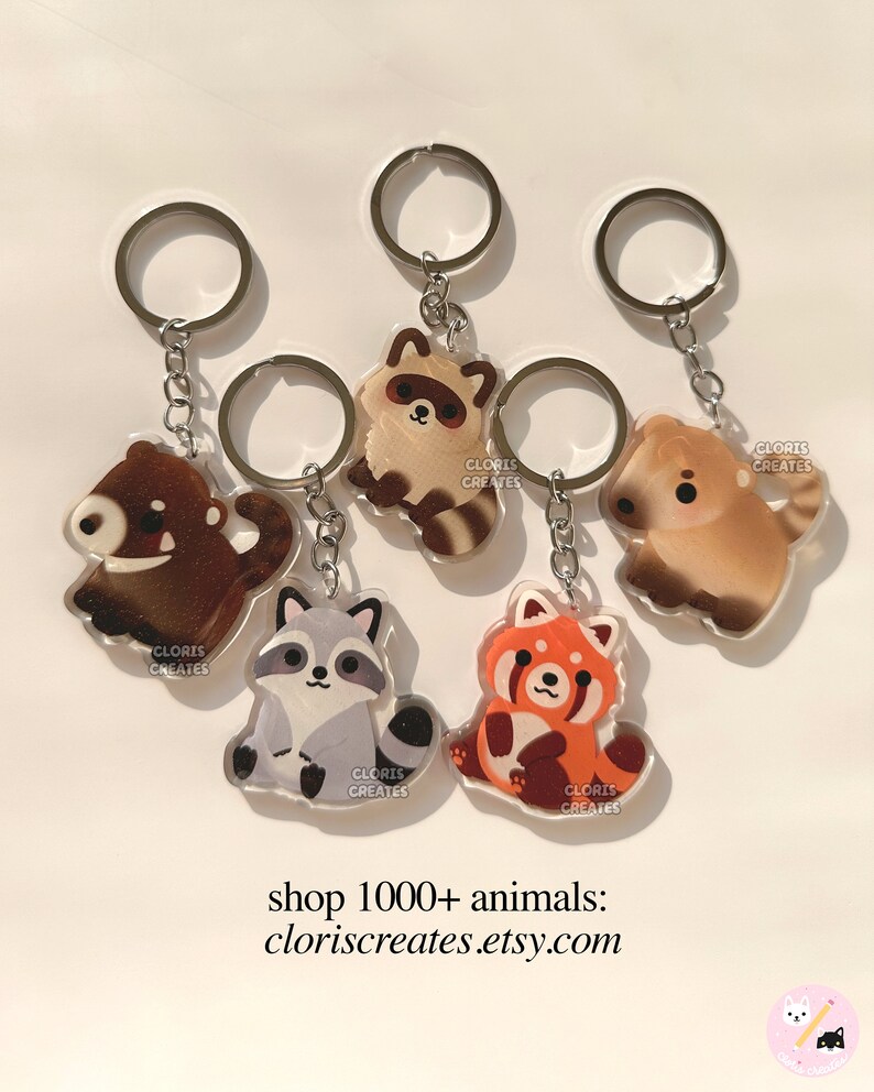 May include: Five different animal keychains with silver key rings. The keychains are made of clear acrylic and feature a raccoon, a red panda, a bear, and two different dogs. Each keychain has the text "CLORIS CREATES" printed on it.