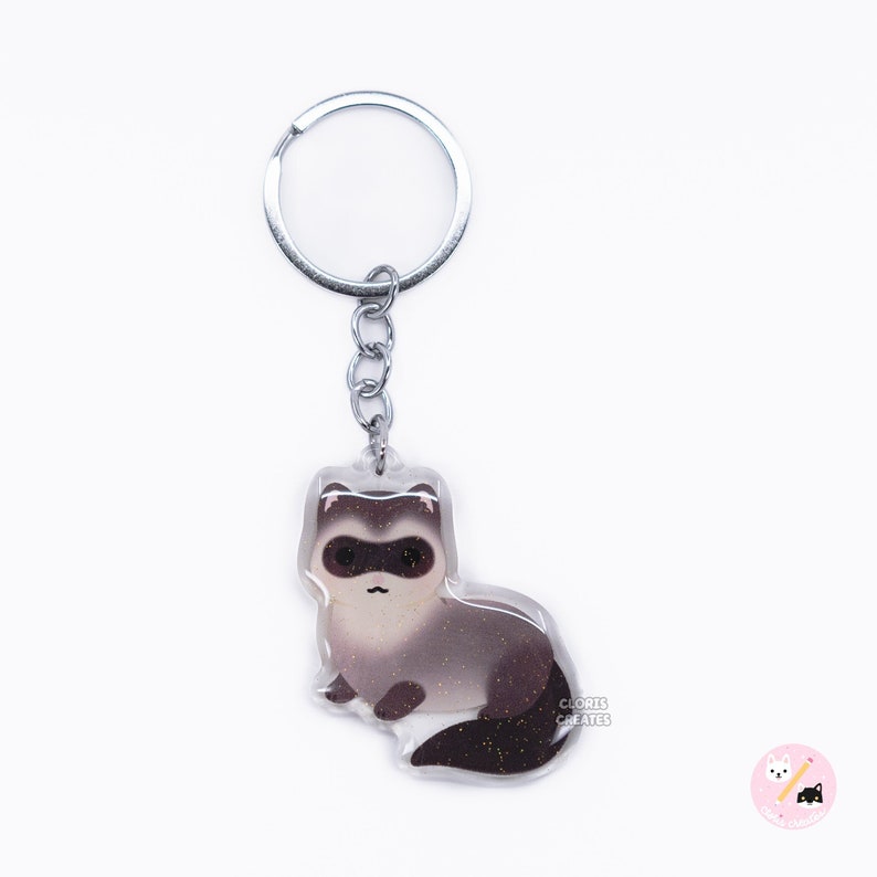 Op de afbeelding: Een zilveren sleutelhanger met een transparante acrylbedel met een cartoon fret met bruin en wit bont en een glinsterende staart. De bedel heeft de tekst "CLORIS CREATES" onderaan gedrukt.