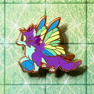 May include: A purple and blue fantasy creature enamel pin with gold accents, holding a blue die. The pin has green and yellow wings. The pin is on a green and white game board with the text "Flare Wizard spell".