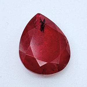 May include: A pear-shaped faceted gemstone with a deep red color. The stone features a pointed top and a rounded base with multiple triangular facets reflecting light. There are visible dark inclusions within the translucent red structure.