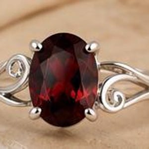 May include: A silver ring featuring a large, oval-cut, deep red garnet gemstone held by four prongs. The band is crafted from polished silver and displays intricate, swirling, open-work metal patterns on both sides of the central stone.