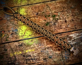 Puede incluir: Un brazalete de paracord negro y marrón con un dije de Mjolnir plateado. El brazalete está tejido con un patrón de nudos y tiene un bucle para sujetarlo a un llavero o una bolsa.