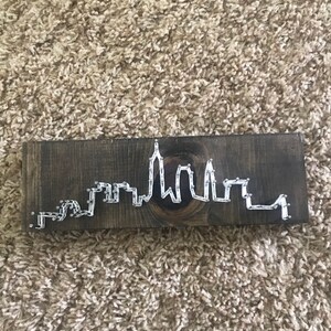 May include: A wooden sign with a white string art design of a city skyline.