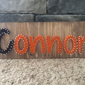 May include: A wooden sign with the name "Connor" spelled out in orange and black string art.