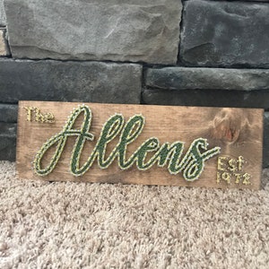 May include: Wooden sign with the name "The Allens" crafted with green and gold string art. The sign also includes "Est 1972" in gold. The sign is mounted on a wooden plank with a rustic finish, perfect for home decor.