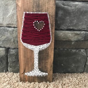 May include: A red wine glass with a white heart in the center, made with string wrapped around nails on a wooden board.