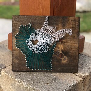 May include: A wooden plaque with a string art design of the state of West Virginia. The state is outlined in white string, and the interior is filled with green string.