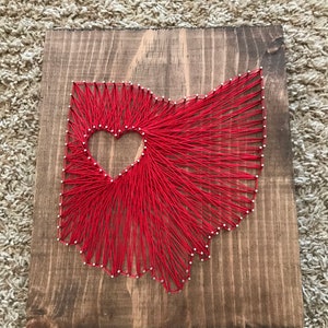 May include: Red string art of the state of Ohio with a heart shape in the middle. The art is on a wooden board.
