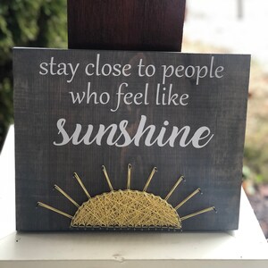 May include: A wooden sign with the text "stay close to people who feel like sunshine" in white lettering. A yellow string art sun is below the text.