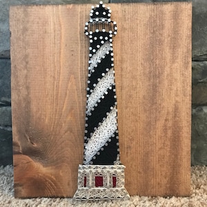May include: A handcrafted string art piece featuring a lighthouse design. The lighthouse is black with white diagonal stripes and a white top, mounted on a wooden board. The base of the lighthouse has red accents.