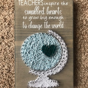 May include: A wooden sign with the text "TEACHERS inspire the smallest hearts to grow big enough to change the world". A string art globe with a teal heart in the center is displayed on the sign.