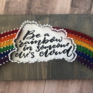 May include: A wooden sign with a rainbow made of string wrapped around nails. The rainbow is in a white cloud with the text "Be a rainbow in someone else's cloud" written in black.