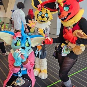 May include: Three full-body animal costumes featuring vibrant colors. One is blue with large ears and a pink flower, one is yellow with glasses, and one is red with a yellow collar. They wear accessories like badges and carry a small plush toy.
