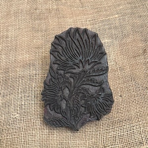 May include: A dark brown, rectangular wooden block with a floral design. The carved pattern features a large central flower with detailed petals and leaves, along with smaller flowers. The block is set on a rough, tan burlap surface.