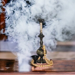 May include: A black metal hookah with a gold beaded hose and a bowl, surrounded by white smoke on a wooden surface.