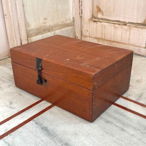 May include: A rectangular wooden storage box with a medium brown finish and visible wood grain. The box features a hinged lid and a black metal latch mechanism on the front center. The surface of the wood shows minor signs of wear and texture.