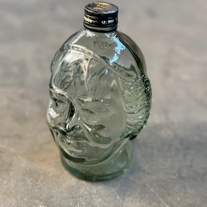 May include: A clear glass bottle shaped like a human head with a black lid. The bottle is green tinted and has a textured surface.