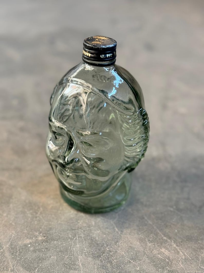 May include: A clear glass bottle shaped like a human head with a black lid. The bottle is green tinted and has a textured surface.