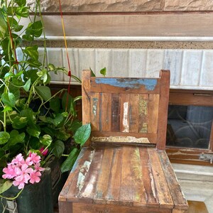 Puede incluir: Una silla de madera r&uacute;stica con un acabado desgastado. La silla tiene un marco de madera marr&oacute;n claro con un respaldo pintado en azul, blanco y amarillo. El asiento est&aacute; hecho de m&uacute;ltiples piezas de madera, creando un aspecto &uacute;nico y texturizado.