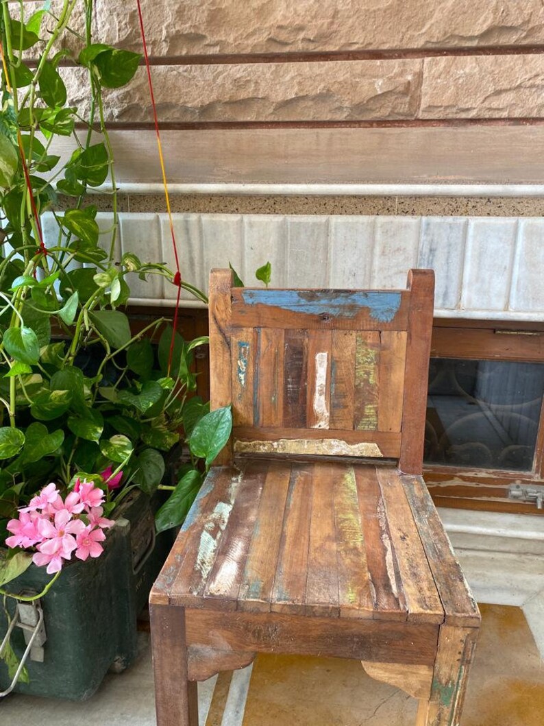 Puede incluir: Una silla de madera r&uacute;stica con un acabado desgastado. La silla tiene un marco de madera marr&oacute;n claro con un respaldo pintado en azul, blanco y amarillo. El asiento est&aacute; hecho de m&uacute;ltiples piezas de madera, creando un aspecto &uacute;nico y texturizado.