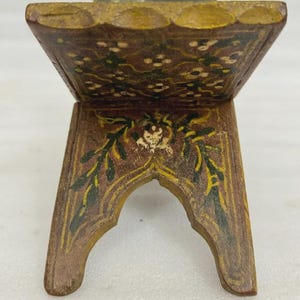 Puede incluir: Un peque&ntilde;o soporte decorativo de madera con tapa con bisagras. El soporte est&aacute; pintado de marr&oacute;n con dise&ntilde;os florales y de hojas en verde, amarillo y blanco. El soporte tiene una base arqueada y probablemente se utiliza para exhibir.