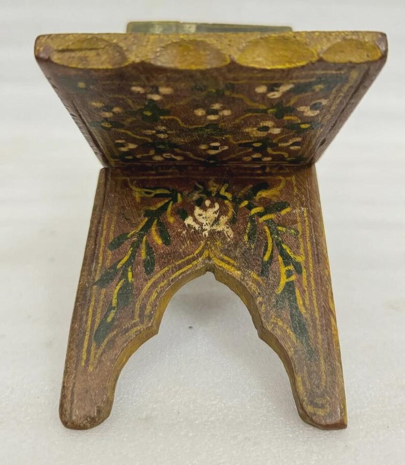 Puede incluir: Un peque&ntilde;o soporte decorativo de madera con tapa con bisagras. El soporte est&aacute; pintado de marr&oacute;n con dise&ntilde;os florales y de hojas en verde, amarillo y blanco. El soporte tiene una base arqueada y probablemente se utiliza para exhibir.