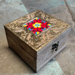 Puede incluir: Caja de madera cuadrada con marco decorativo de latón e incrustaciones de azulejos florales de colores. La caja tiene una tapa con bisagras y un cierre metálico. La madera tiene un acabado natural marrón claro.
