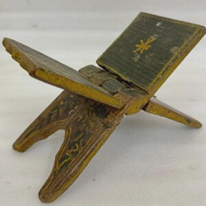 Puede incluir: Un peque&ntilde;o atril de madera antiguo con un dise&ntilde;o de bisagra. Pintado en tonos verdes, amarillos y marrones, con detalles florales. La secci&oacute;n superior presenta un dise&ntilde;o floral amarillo. El atril est&aacute; dise&ntilde;ado para sostener un libro abierto.