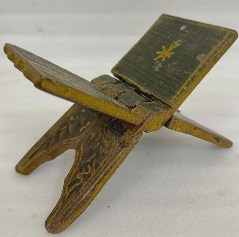 Puede incluir: Un peque&ntilde;o atril de madera antiguo con un dise&ntilde;o de bisagra. Pintado en tonos verdes, amarillos y marrones, con detalles florales. La secci&oacute;n superior presenta un dise&ntilde;o floral amarillo. El atril est&aacute; dise&ntilde;ado para sostener un libro abierto.